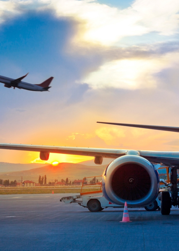 Ein Flugzeug am Himmel über einem geparkten Flugzeug am Flughafen vor einer hügeligen Landschaft bei Sonnenuntergang.