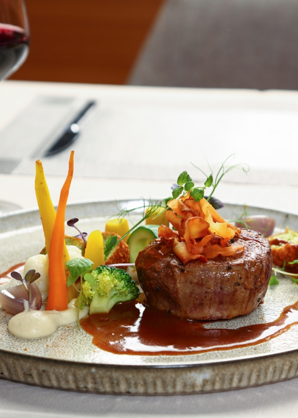 Ein Stück Steak an Gemüse, angerichtet auf einem Teller neben einem Glas Rotwein auf einem weiß gedeckten Tisch.