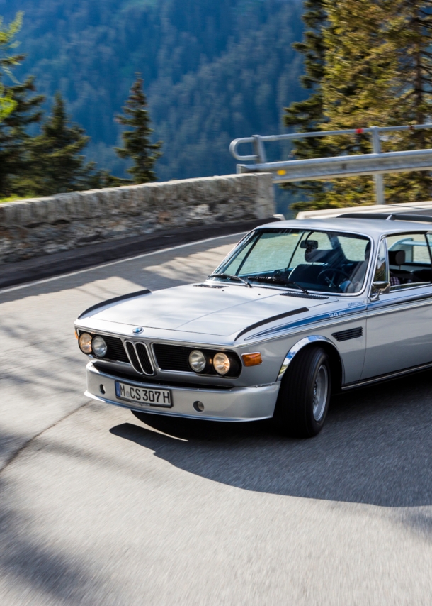 Ein fahrender silberner BMW 3.0 CSL in einer Kurve auf einer Straße in den Bergen.