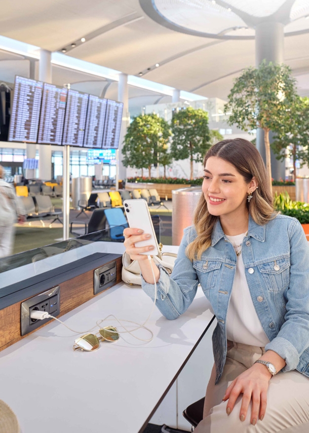 Eine junge Frau mit Smartphone sitzt in einem Wartebereich am Flughafen.