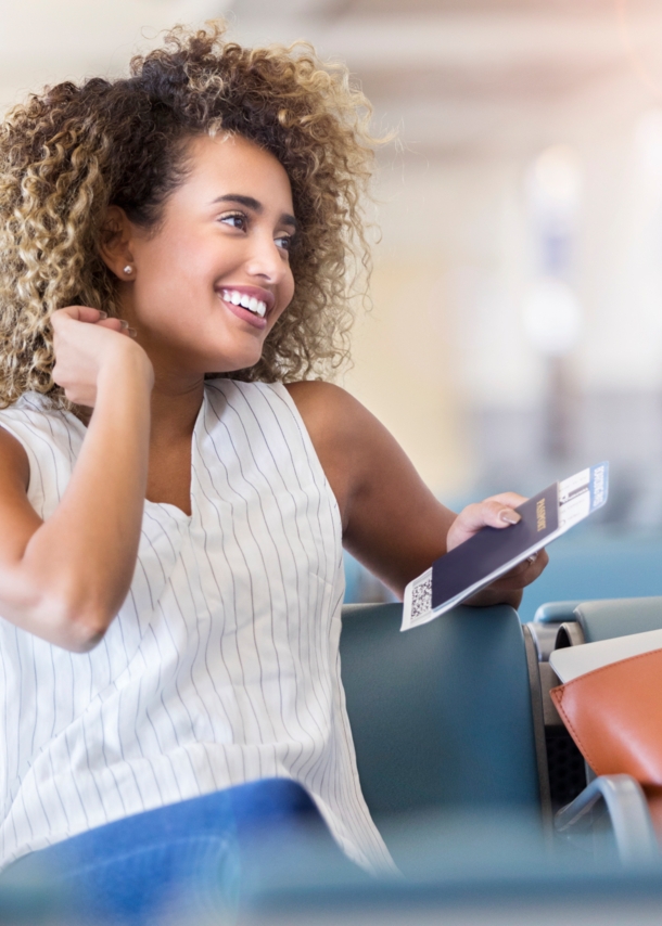 Eine junge, lächelnde Frau sitz mit Reisepass in der Hand in einer Flughafenlounge.