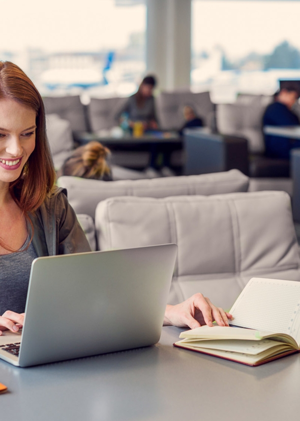 Eine lächelnde Frau arbeitet an ihrem Laptop an einem Tisch in einer Flughafen-Lounge.