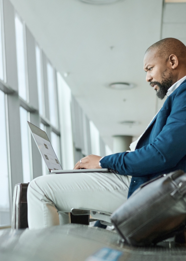 Ein Mann, der am Flughafen am Fenster sitzt und einen aufgeklappten Laptop auf dem Schoß hat.