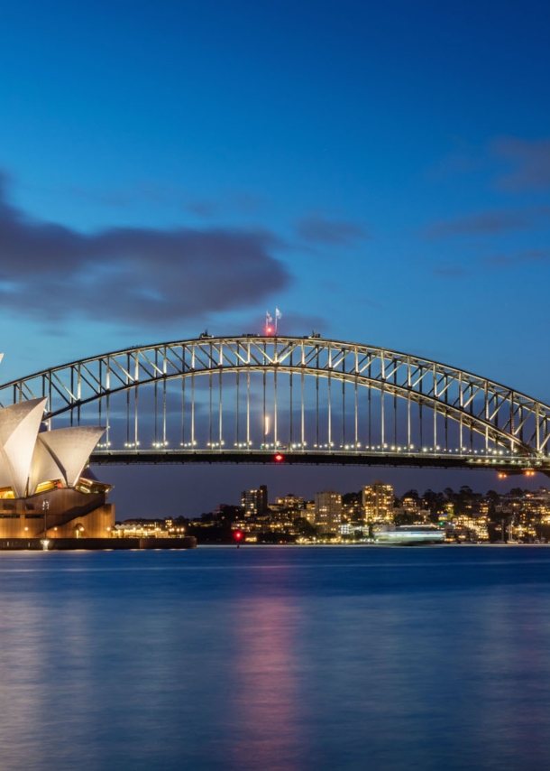 Sydney Harbour Bogenbrücke hinter dem Sydney Opernhaus am Wasser vor der erleuchteten Skyline Sydneys bei Nacht.