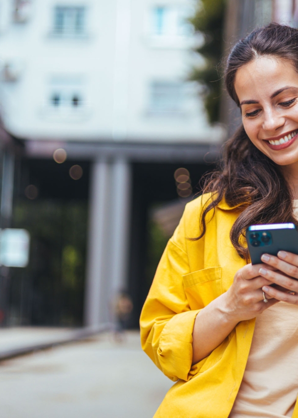 Eine Frau im gelben Hemd lehnt an einer Außenfassade und schaut lächelnd auf das Smartphone in ihren Händen.