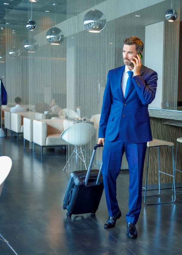 Ein Geschäftsmann im blauen Anzug und mit Trolley telefoniert stehend in einer modernen Flughafen-Lounge.