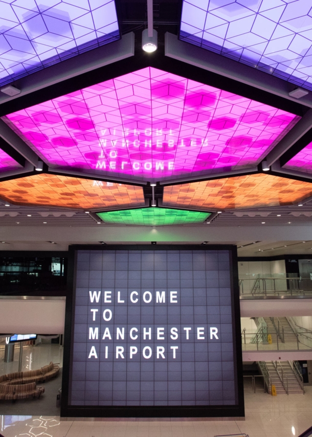 Menschenleere Flughafenhalle mit einem Bildschirm im Mittelpunkt, auf dem „Welcome to Manchester Airport“ steht und bunt erleuchteten LED-panels an der Decke.