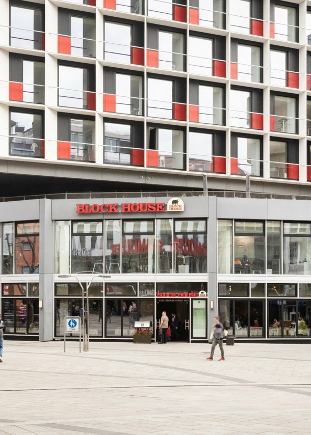 Personen am Eingang einer Block House Filiale in einem grauen Neubau in einer Fußgängerzone in Innenstadtlage.