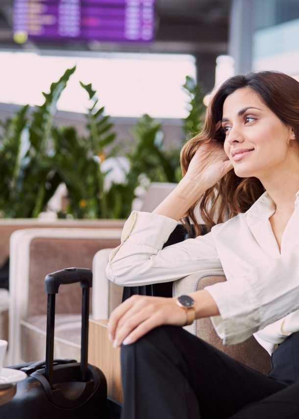 Eine Frau in Business-Kleidung sitzt in einem hellen Lounge-Sessel. Neben ihr steht ein Koffer, auf dem Tisch vor ihr befinden sich Reisedokumente und eine Tasse mit Untersetzer.