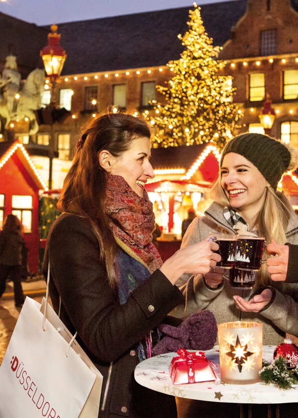 Drei Personen mit Glühweintassen an einem Tisch auf einem Weihnachtsmarkt am Düsseldorfer Rathaus.