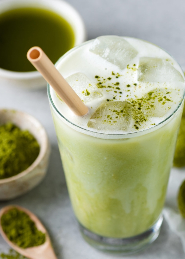 Ein Glas mit Matcha Latte und Strohhalm, daneben liegen grüne Kugeln, eine Schale mit grünem Pulver, ein Bambusbesen und eine Schale mit purem Matchatee.