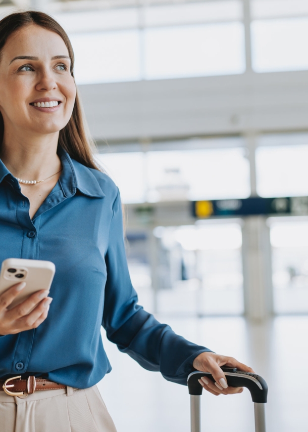 Eine Frau am Flughafen, die in einer Hand einen Koffergriff hält und in der anderen ein Smartphone.