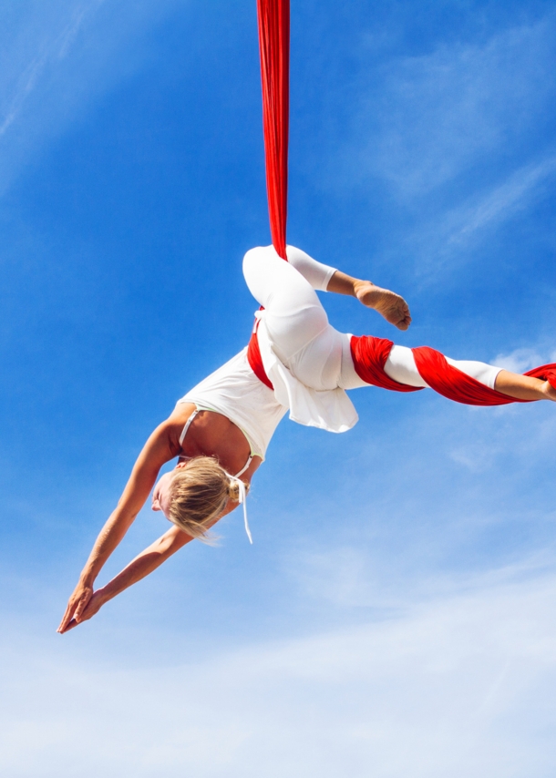 Untersicht einer Frau in weißer Kleidung in schwebender Yoga-Pose in einem roten Tuch vor blauem Himmel.