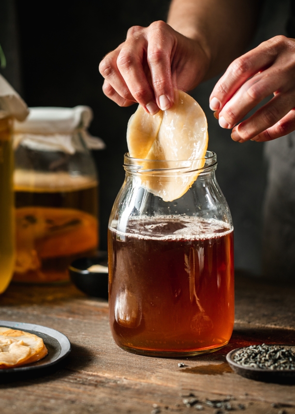 Ein Kombucha-Pilz wird von einer Hand in ein Gefäß mit brauner Flüssigkeit auf einem Holztisch gegeben, daneben weitere befüllte Einmachgläser.