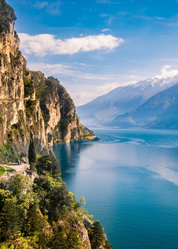 Bewachsene Felswand nahe Riva del Garda direkt am Gardasee mit Gipfeln auf der anderen Seeseite im Hintergrund.