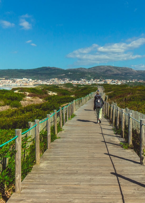 Eine unkenntliche Person beim Wandern in Portugal auf einem Holzsteg, der in Küstennähe über grüne Vegetation führt.