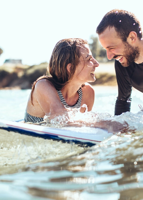 Ein lachendes Paaar mit Surfboard im Meer.