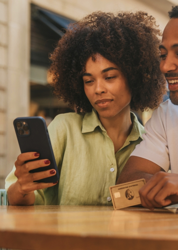 Ein Paar steht an einem Tisch auf einer Einkaufsstraße und schaut gemeinsam auf ein Smartphone, er hält eine goldene Kreditkarte von American Express in der Hand.