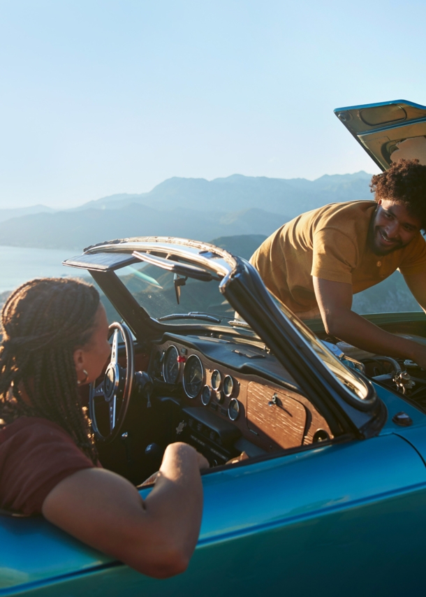 Ein Paar in einem liegengebliebenen Cabriolet an einer Meeresküste bei Sonnenschein, er kontrolliert den Motor, sie sitzt auf dem Beifahrersitz.