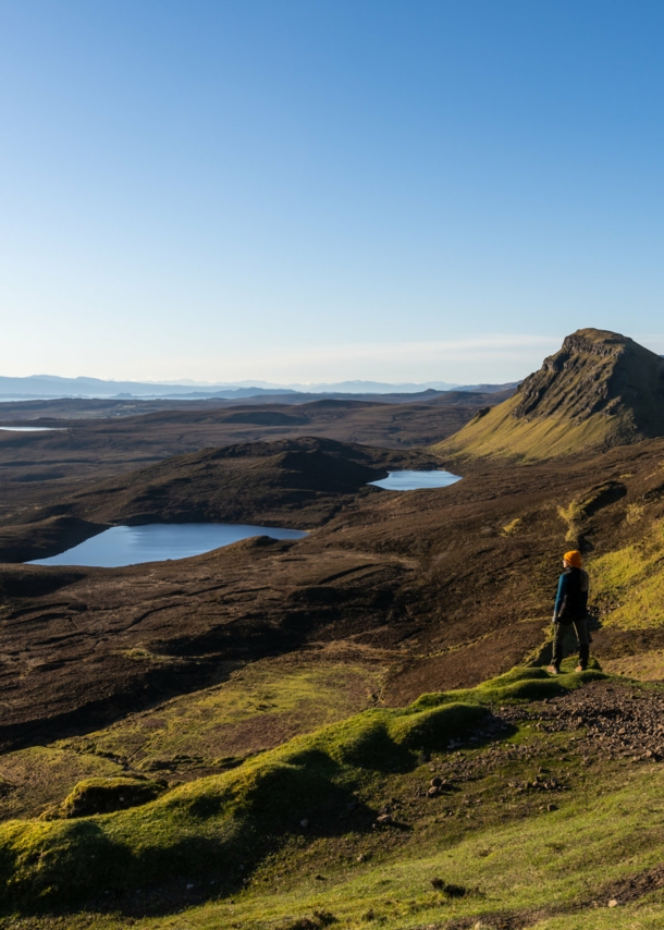 Eine Person steht in einer Landschaft aus Wiesen, Hügeln und Seen in Schottland. 