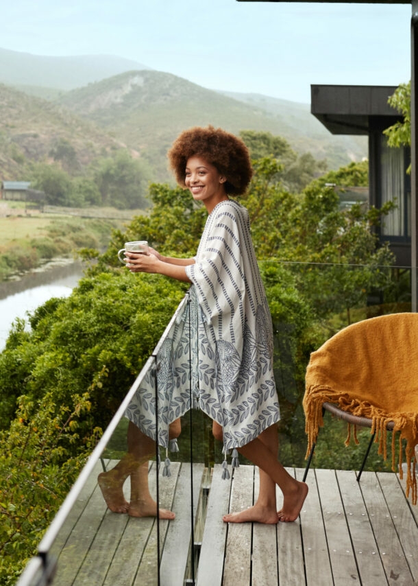 Eine lächelnde Frau steht mit einer Kaffeetasse auf dem Balkon eines Luxushotels mit Blick auf eine grüne Buschlandschaft mit Fluss.