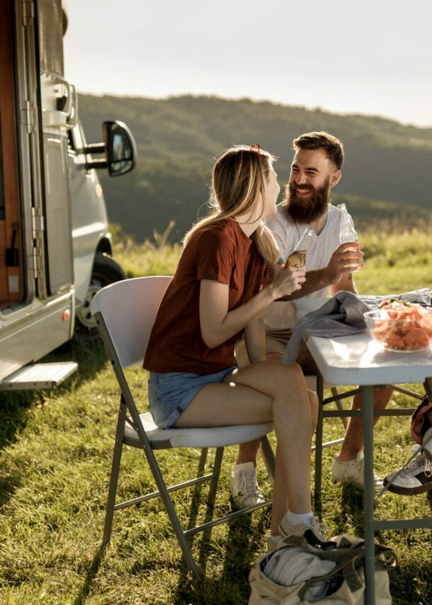 Vier gut gelaunte Personen in Sommerkleidung sitzen an einem Campingtisch neben einem Wohnmobil auf einer Rasenfläche vor Hügellandschaft.