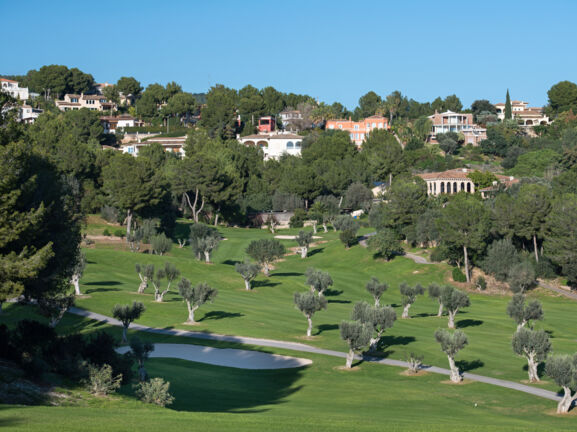 Ausschnitt des Golfklubs mit einem Bunker und vielen kleinen Olivenbäumen, vor der Kulisse des Ortes Bendinat