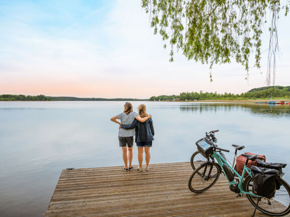 Rückansicht von zwei Frauen, die auf einem Steg vor einem See stehen und sich umarmen, daneben zwei E-Bikes