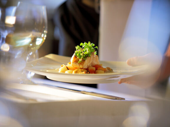 Nahaufnahme eines edel angerichteten Fischgerichtes, das auf einem weißen Teller an einem Tisch mit weißer Tischdecke serviert wird.