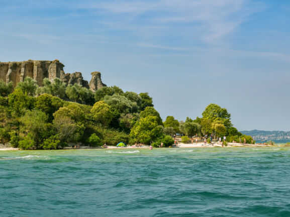 Eine antike Ruine, umgeben von üppiger Natur, an einem Sandstrand mit Personen am türkisblauen Wasser.