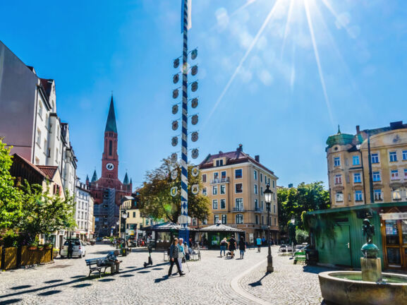 Ein Stadtteilplatz mit Kopfsteinpflaster und einem Maibaum in der Mitte bei strahlendem Sonnenschein.