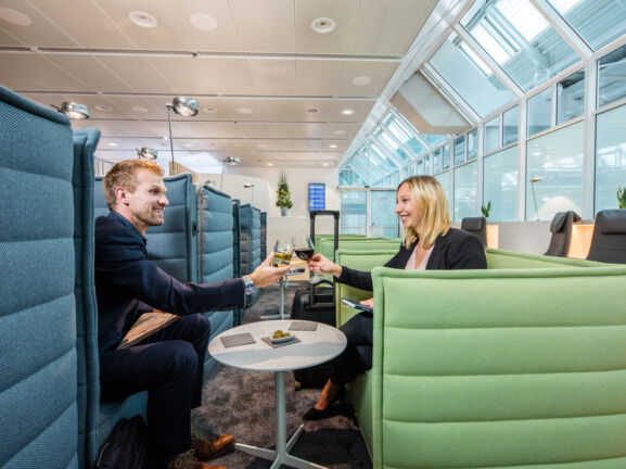 Ein Mann und eine Frau sitzen sich gegenüber auf Sesseln in einer Flughafenlounge und stoßen mit Weingläsern an.