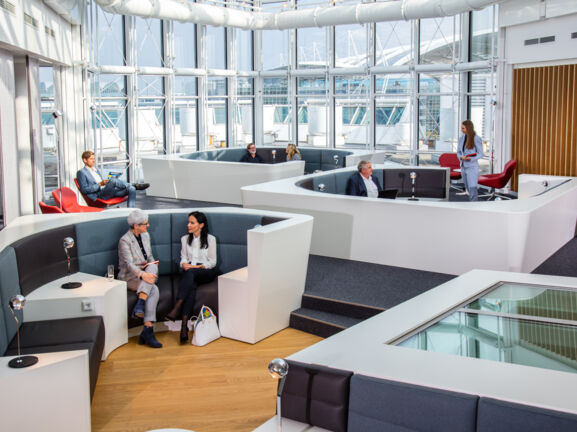 Mehrere Personen sitzen in einer modernen Sitzlandschaft einer Flughafenlounge mit Panoramafenstern.