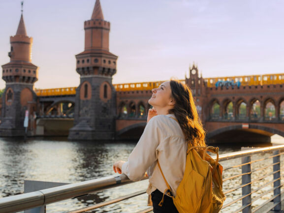 Eine junge Frau mit Rucksack steht am Wasser vor einer Brücke mit Türmen, auf der eine gelbe Straßenbahn fährt.