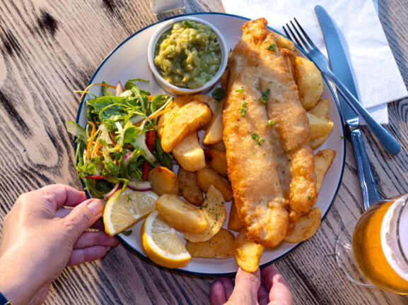 Eine Hand greift nach einem Teller Fish and Chips mit Beilagen auf einem Holztisch bei Sonnenschein.