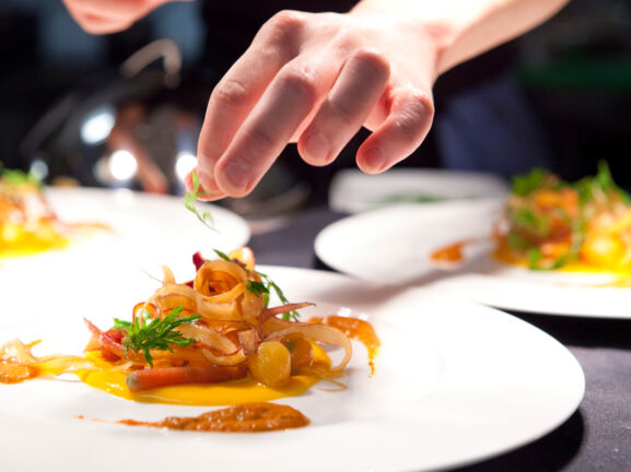 Eine Hand garniert ein Gericht auf einem weißen Teller in einer Restaurantküche.