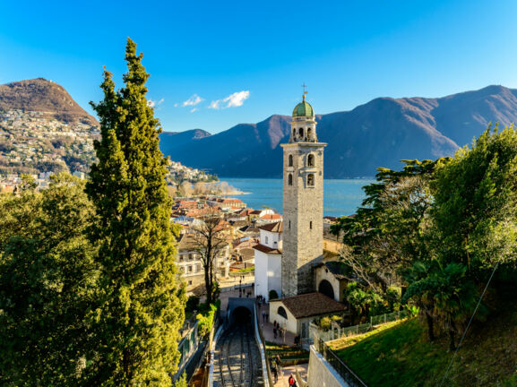 Stadtpanorama von Merano mit Zypressen und Glockenturm an einem See vor Bergpanorama.