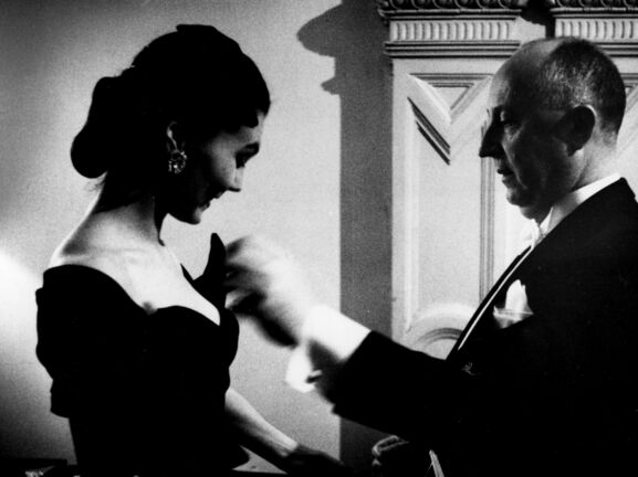 Profilansicht von Christian Dior in einem schwarzen Anzug, der etwas an einem schwarzen Abendkleid eines weiblichen Models stylt, Schwarzweißfoto.