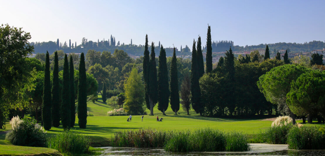 Ein kleiner See mit Blick auf eine Golfbahn und zwei Golfspieler:innen