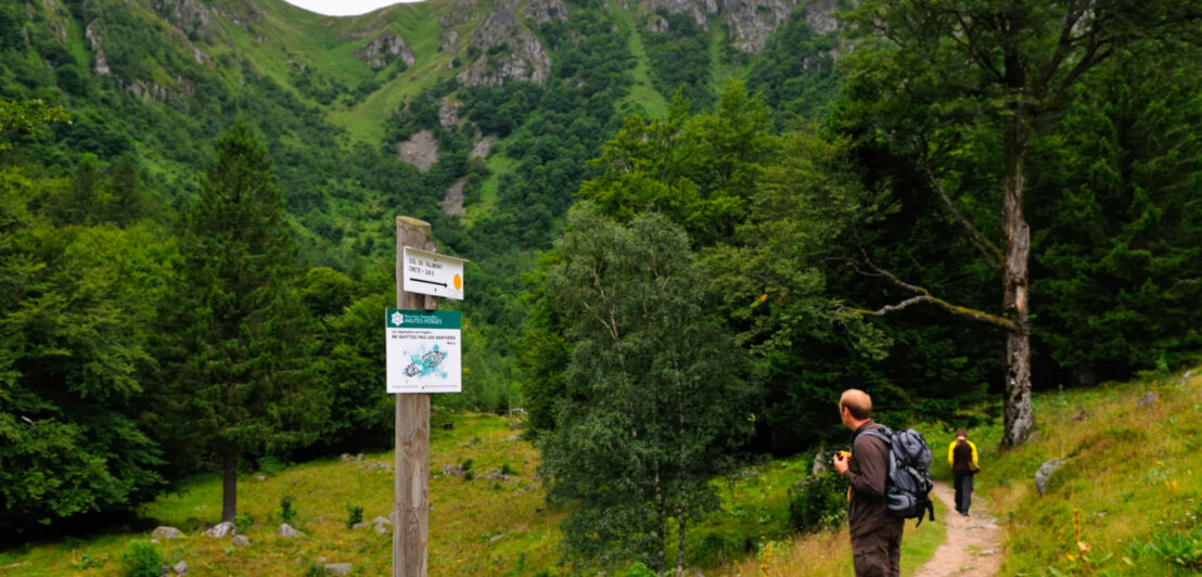 Ein Mann steht vor einem Wegweiser im Hintergrund sind Bäume und Felsen