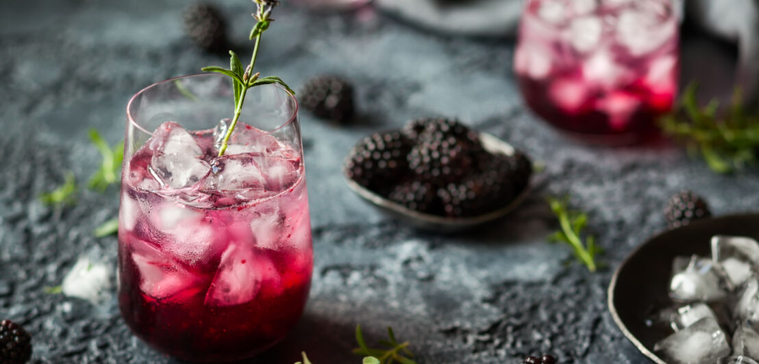 Ein Glas mit einem rötlich gefärbten Longdrink, Kräutern und Brombeeren