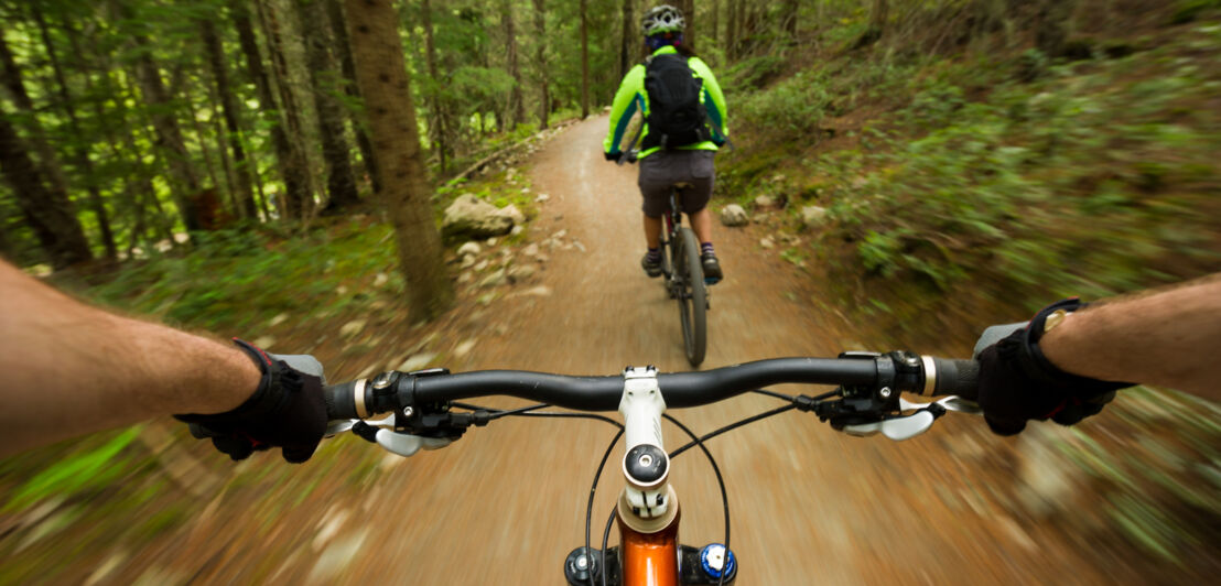 Blick über den Lenker eines Mountainbikes auf eine voranfahrende Mountainbikerin im Wald