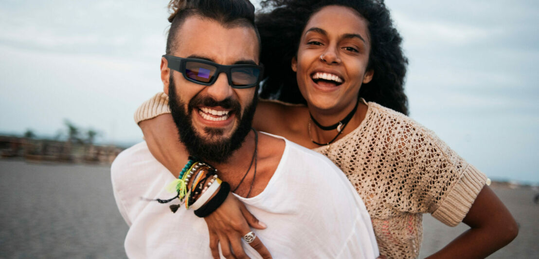 Ein Paar lachend am Strand, er trägt eine Sonnenbrille