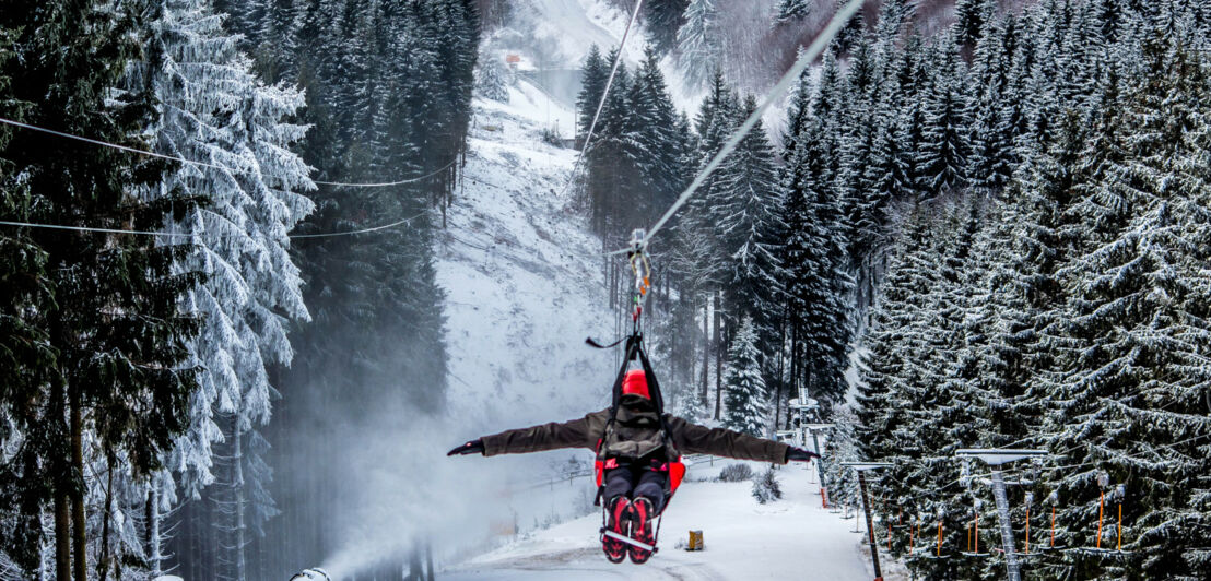 Eine Person in Winterkleidung überquert an der Zipline ein schneebedecktes Waldgebiet