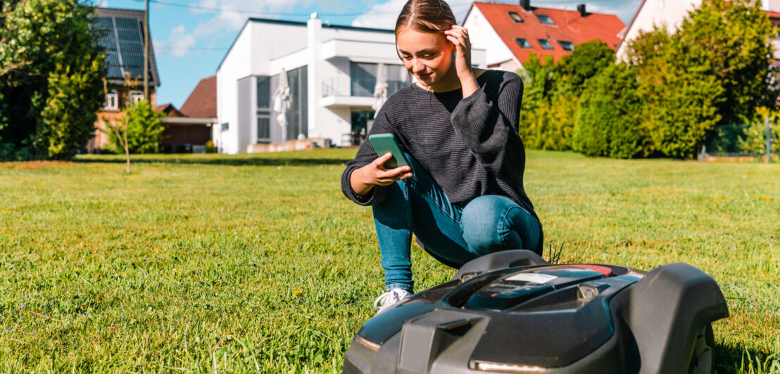 Eine Frau mit Smartphone hockt neben einem Mähroboter auf einer Rasenfläche im Garten