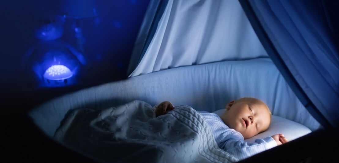 Ein Baby schläft im beleuchteten Babybett in einem dunklen Raum, im Hintergrund blaues Lichtspiel an der Wand