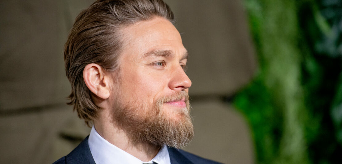 Porträt von Charlie Hunnam mit Vollbart und Anzug vor beige-grünem Hintergrund.