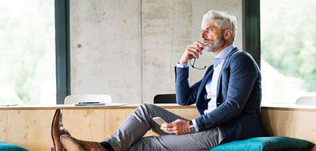 Ein Mann in Businesskleidung sitzt auf einer Holzbank, in den Händen hält er ein Smartphone und eine Brille