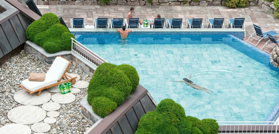 Begrünte, auf Terrassen angelegte Poollandschaft mit Liegestühlen und Personen im und am Wasser aus der Vogelperspektive