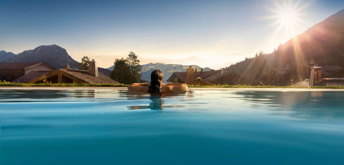 Rückansicht einer Frau im Infinitypool mit Blick über den Ort auf die Berge bei Sonnenschein